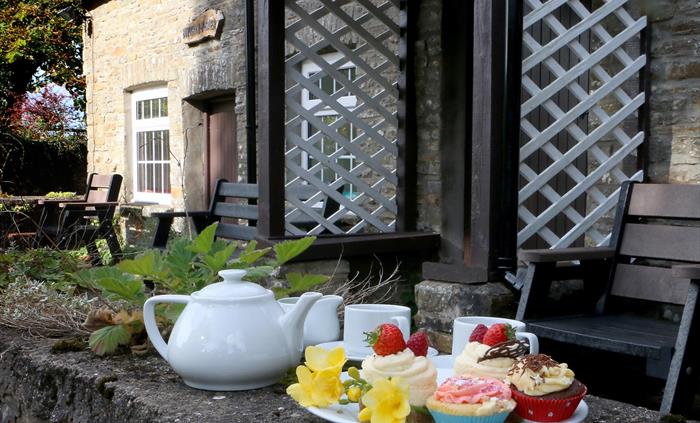 A plate of colourful cupcakes and a white teapot set on a stone wall outside a rustic stone cottage with outdoor seating and trellis.